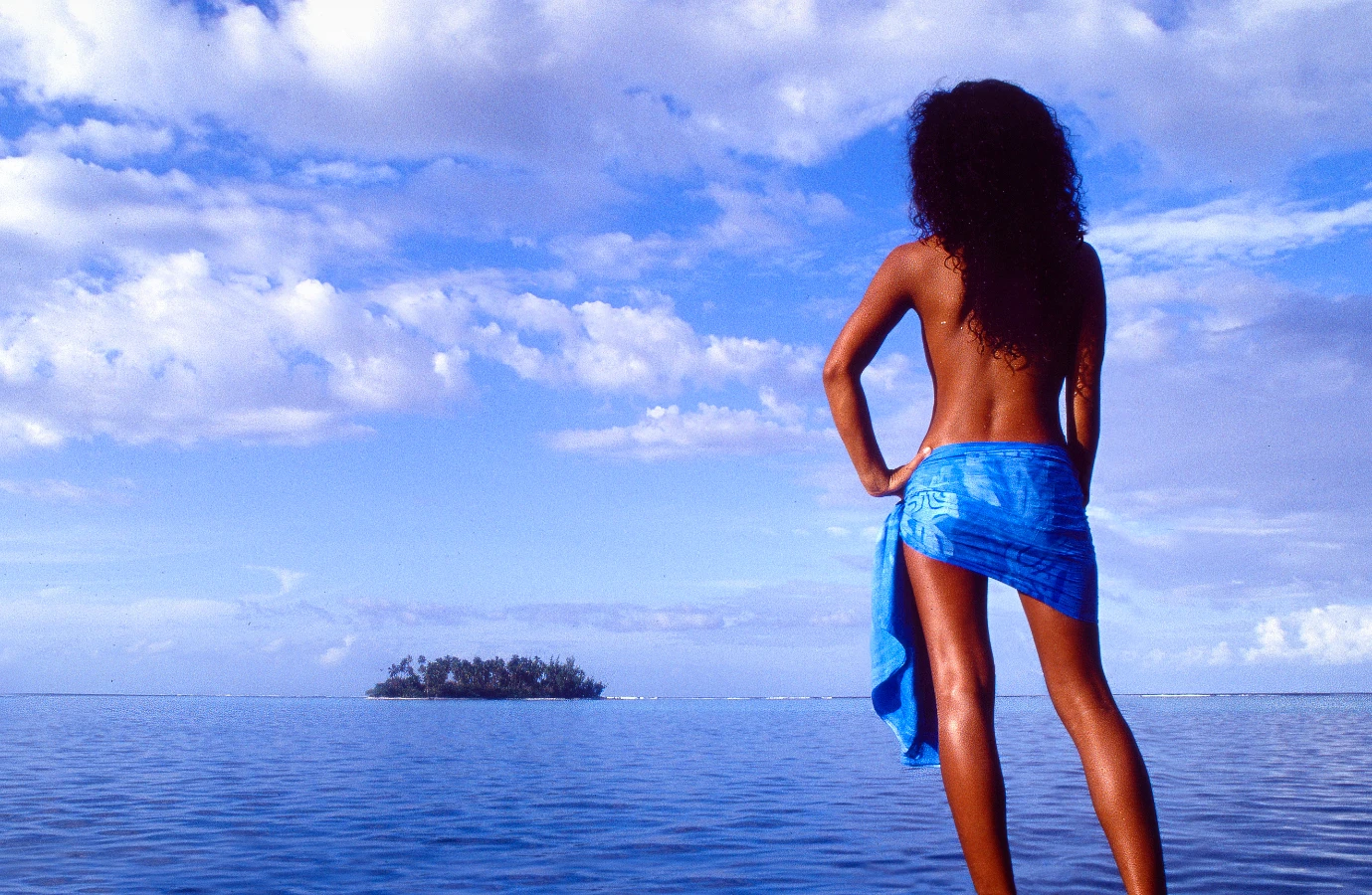 A beautiful Tahitian woman wearing a pareo looks across the ocean.